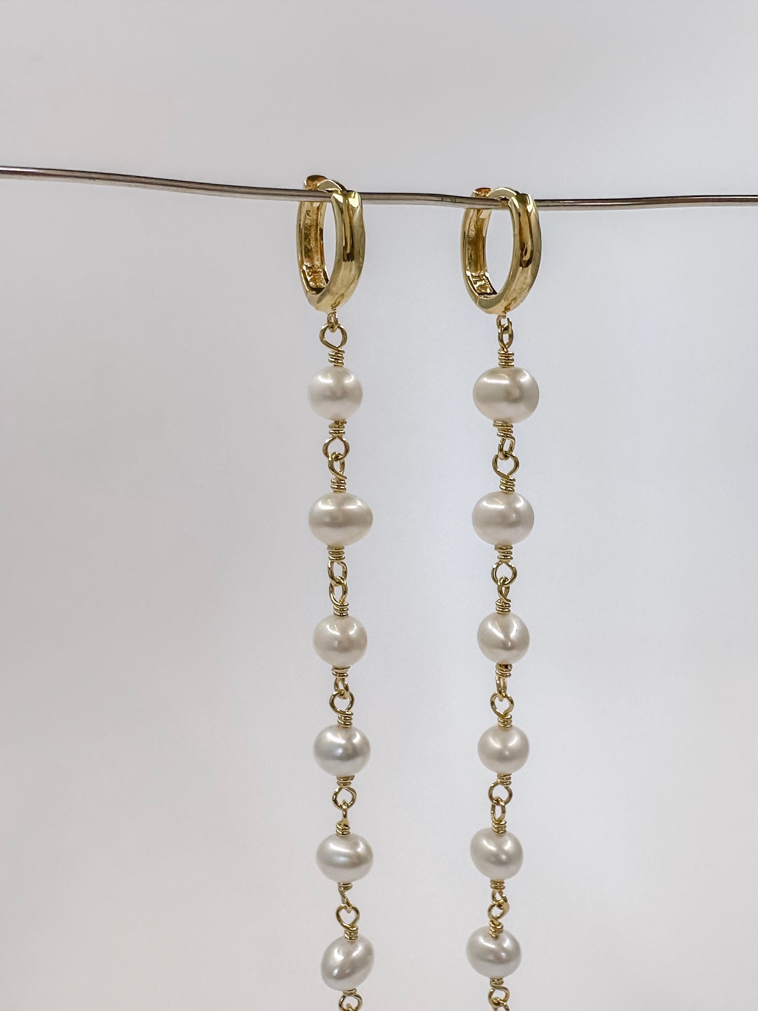 Brinco feito a mão com prata em banho de ouro e 9 pérolas em cada pé finalizando com uma plaquinha na ponta e argola em click pendurado em um fio de metal e fundo branco.