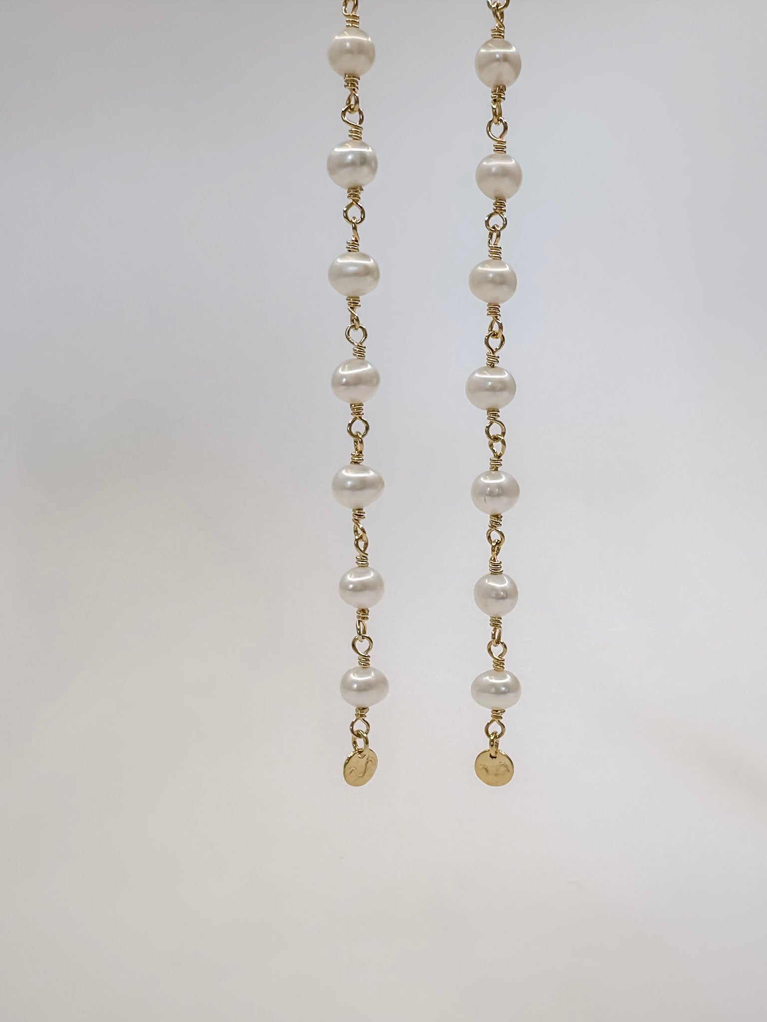 Brinco feito a mão com prata em banho de ouro e 9 pérolas em cada pé finalizando com uma plaquinha na ponta e argola em click pendurado em um fio de metal e fundo branco.