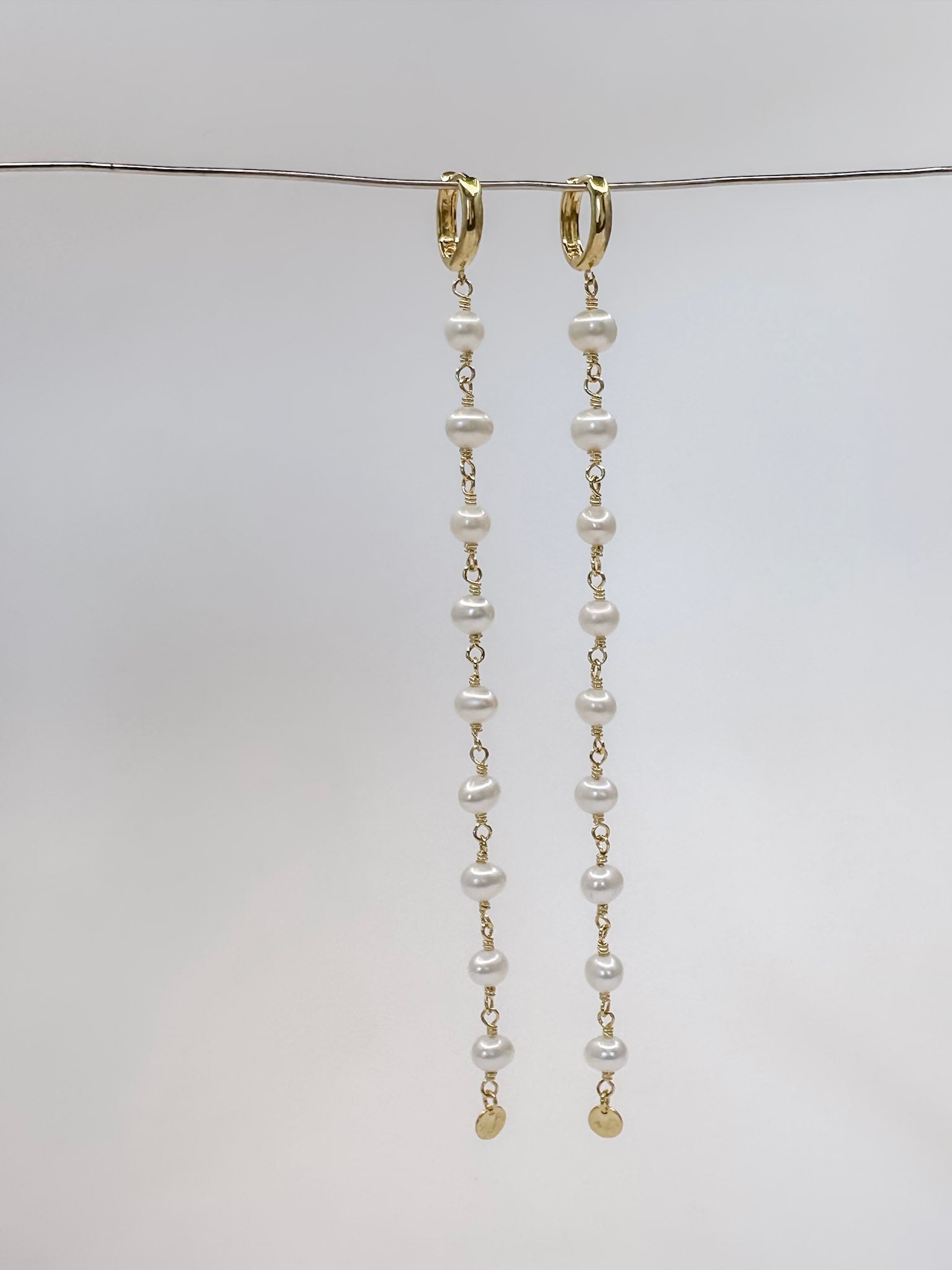 Brinco feito a mão com prata em banho de ouro e 9 pérolas em cada pé finalizando com uma plaquinha na ponta e argola em click pendurado em um fio de metal e fundo branco.