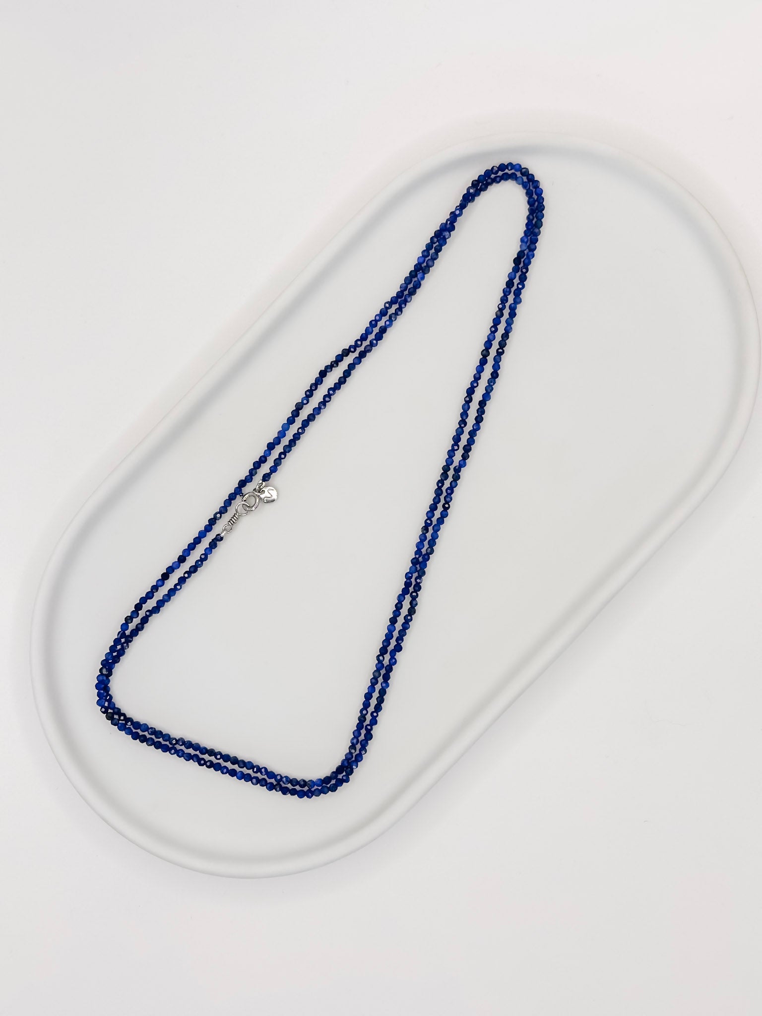 Colar Rasa produzido com esferas lapidadas de pedra natural lapis lazuli de aproximadamente 2 mm com cumprimento de 80 cm e fecho boia em prata com banho de ouro apoiado em bandeja branca de ceramica com fundo branco.