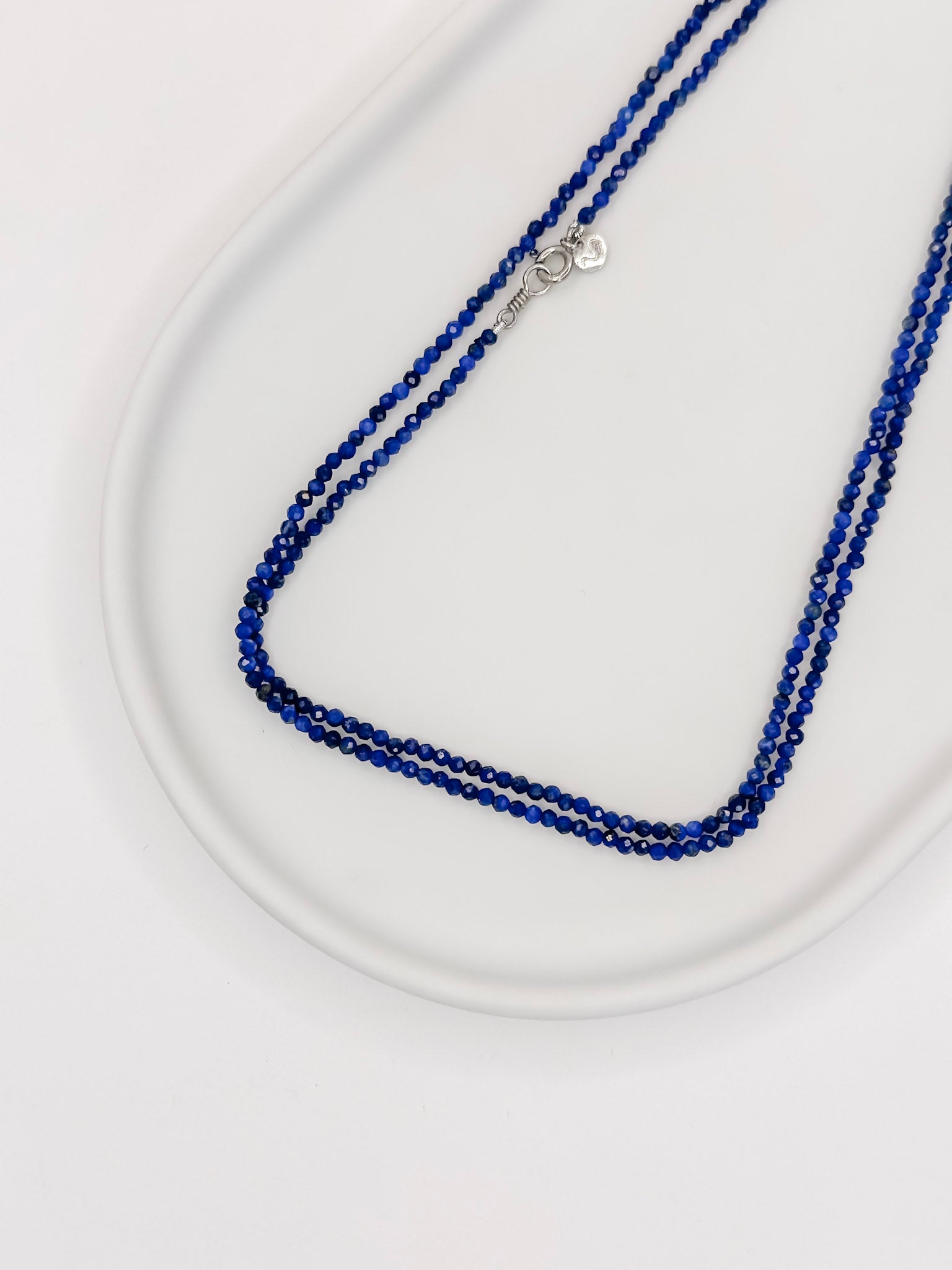 Colar Rasa produzido com esferas lapidadas de pedra natural lapis lazuli de aproximadamente 2 mm com cumprimento de 80 cm e fecho boia em prata com banho de ouro apoiado em bandeja branca de ceramica com fundo branco.