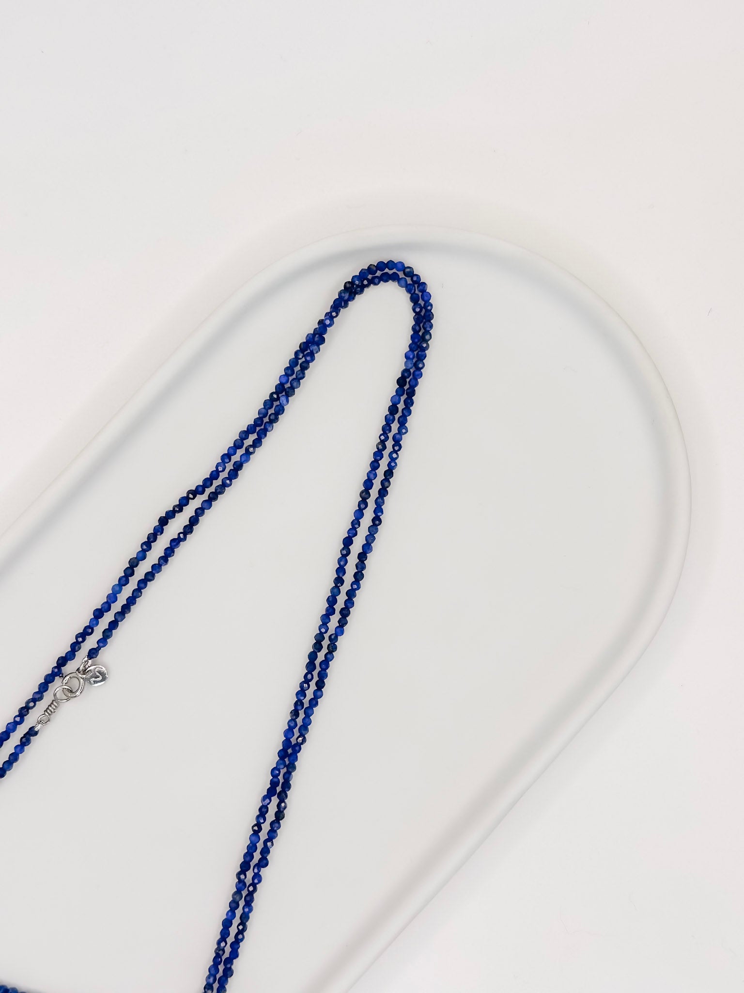 Colar Rasa produzido com esferas lapidadas de pedra natural lapis lazuli de aproximadamente 2 mm com cumprimento de 80 cm e fecho boia em prata com banho de ouro apoiado em bandeja branca de ceramica com fundo branco.