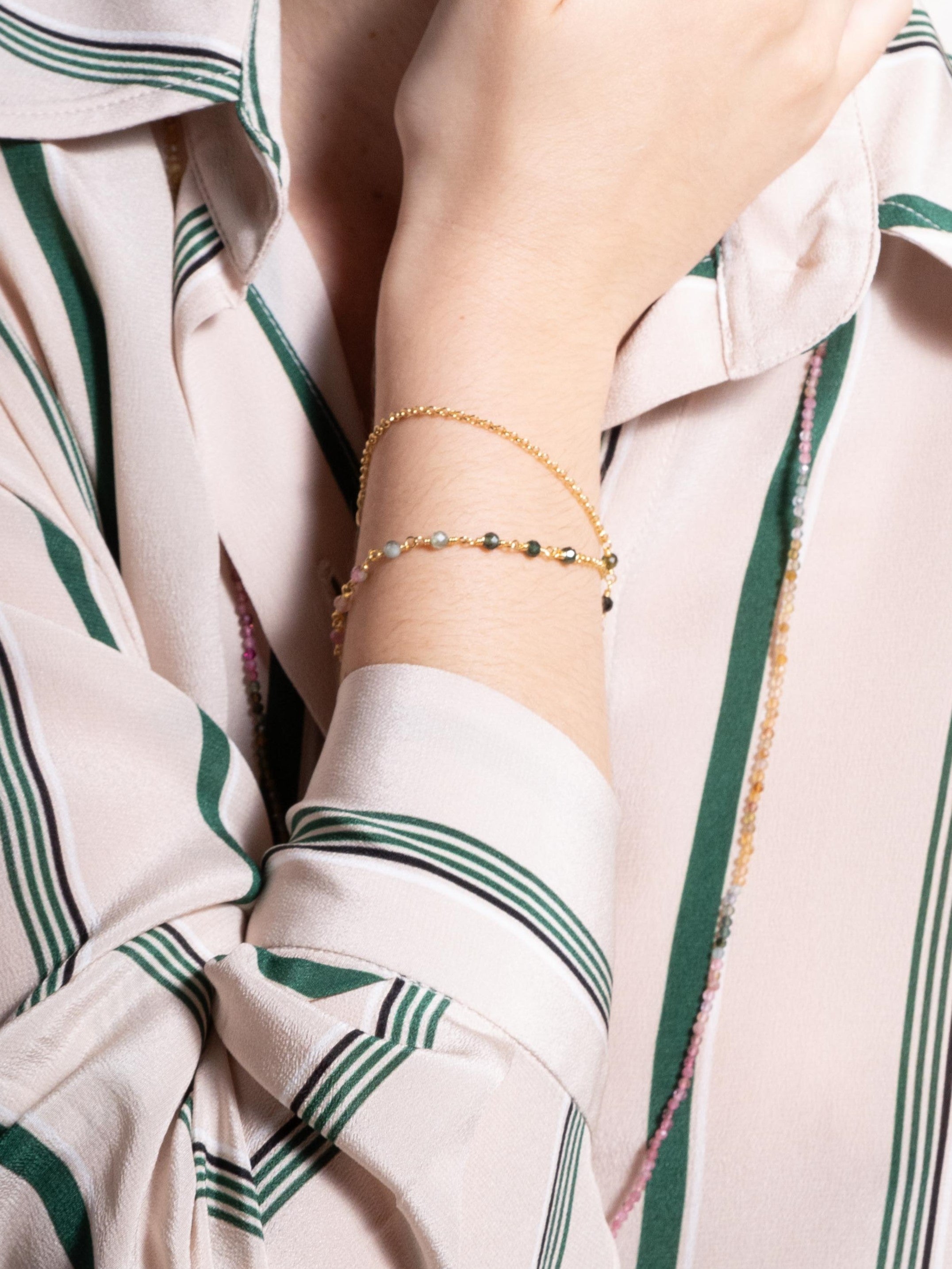 Modelo de pele branca vestindo Pulseira de duas voltas com turmalinas melancia em degrade de cores e corrente portuguesa em prata com banho de ouro no pulso e camisa listrada em tons bege e verde no fundo.
