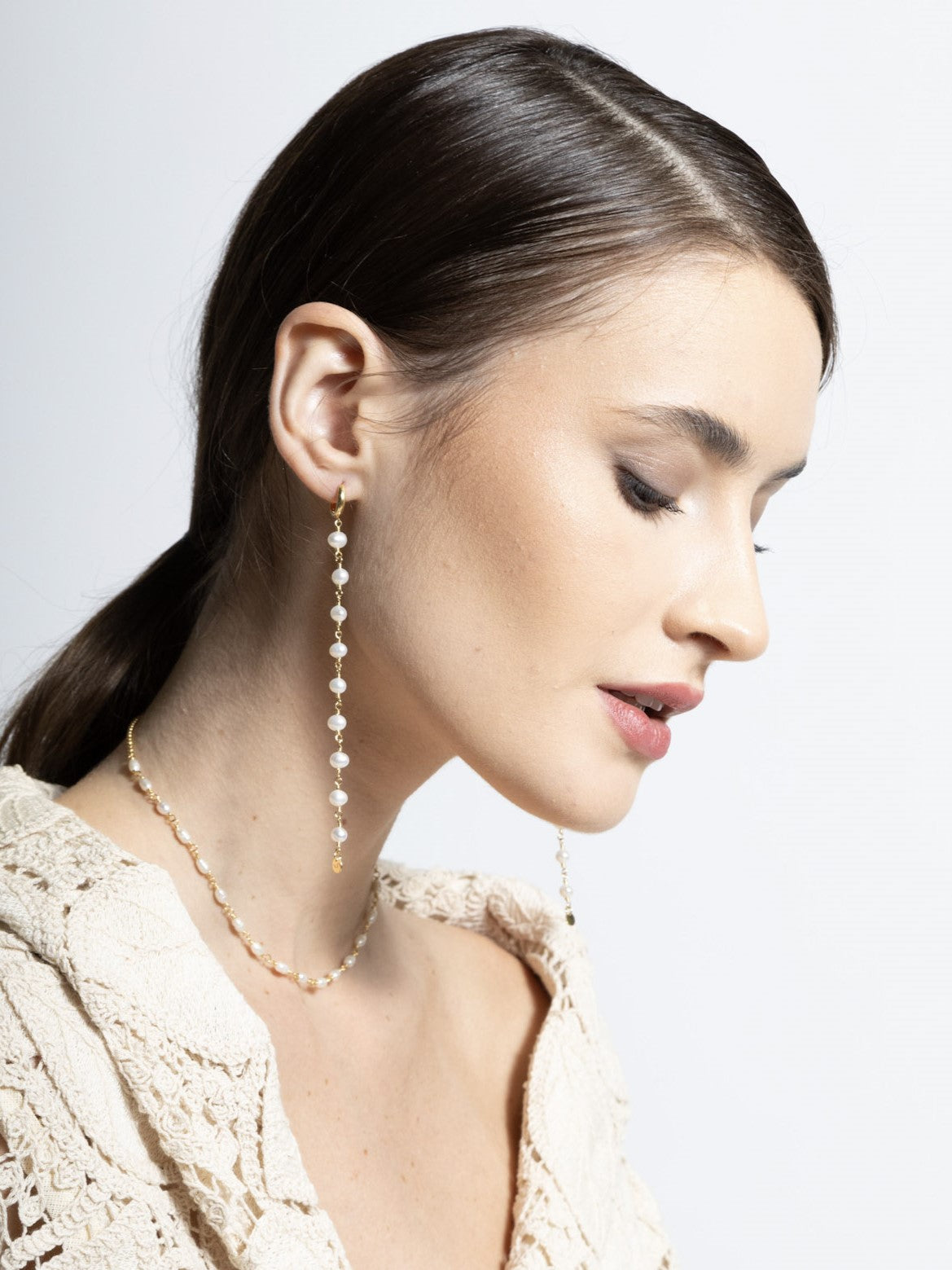 Modelo branca vestindo um blazer de renda e um brinco de perolas longo e colar com perolas de agua doce em prata com banho de ouro. 