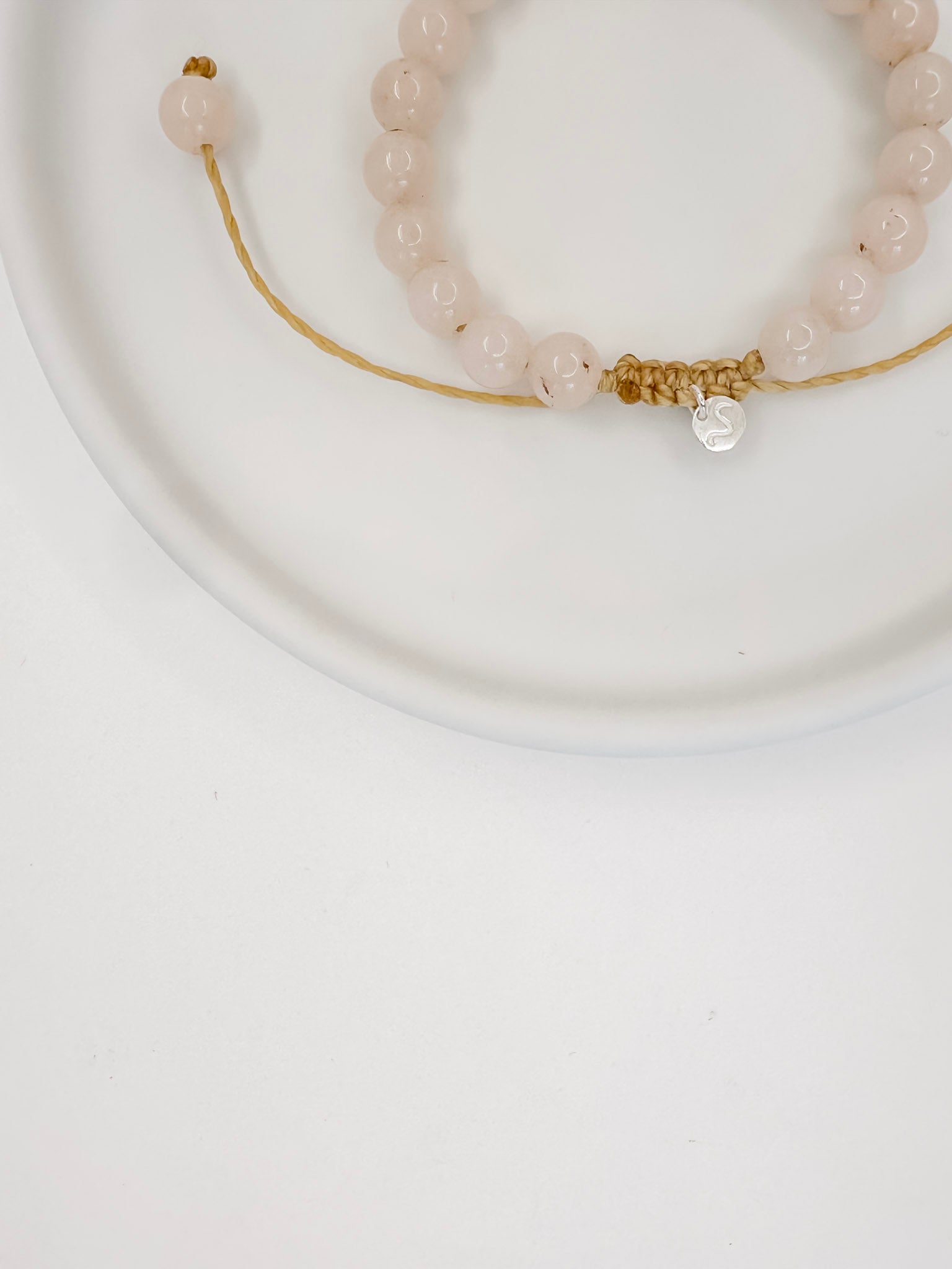 Pulseira feita com corda e esferas lisas de 6mm de cristal quartzo rosa, tamanho unico, apoiada em ceramica branca com fundo branco.