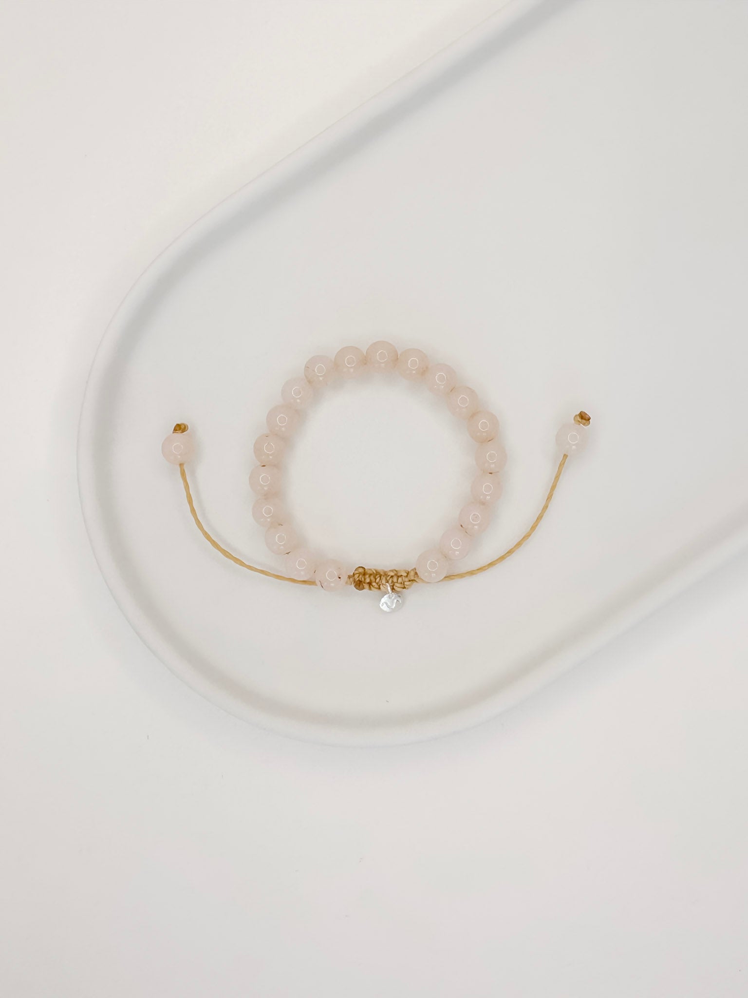 Pulseira feita com corda e esferas lisas de 6mm de cristal quartzo rosa, tamanho unico, apoiada em ceramica branca com fundo branco.