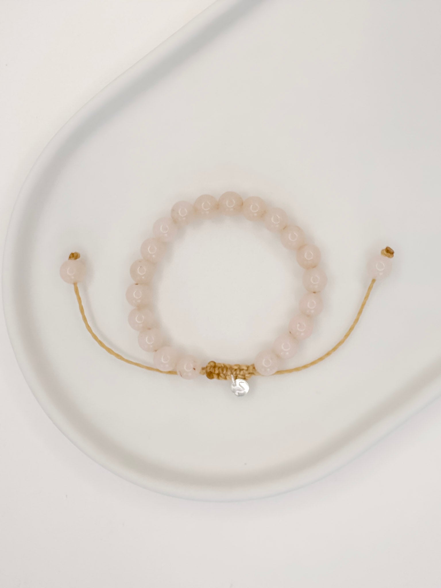 Pulseira feita com corda e esferas lisas de 6mm de cristal quartzo rosa, tamanho unico, apoiada em ceramica branca com fundo branco.