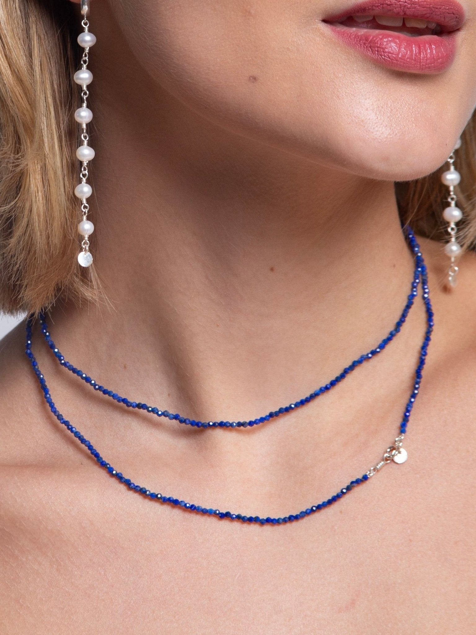 Modelo de pele branca usando Colar Rasa produzido com esferas lapidadas de pedra natural lapis lazuli de aproximadamente 2 mm com cumprimento de 80 cm e fecho boia em prata dando duas voltas no pescoço e brincos longos com perolas naturais.