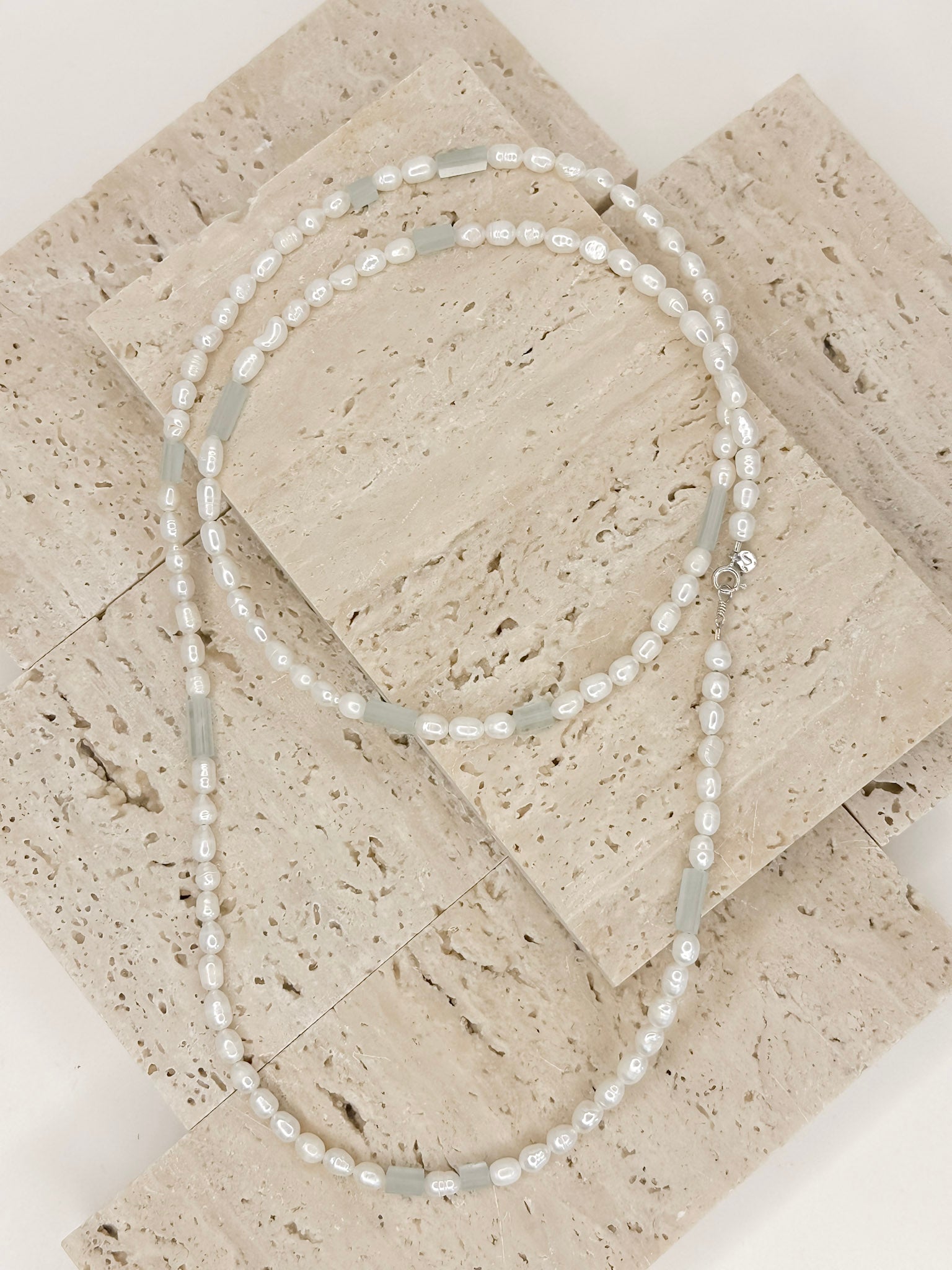 Colar longo com perolas ovais intercalado com pedras naturais água marinha brasileiras, apoiado em marmore com fundo branco.