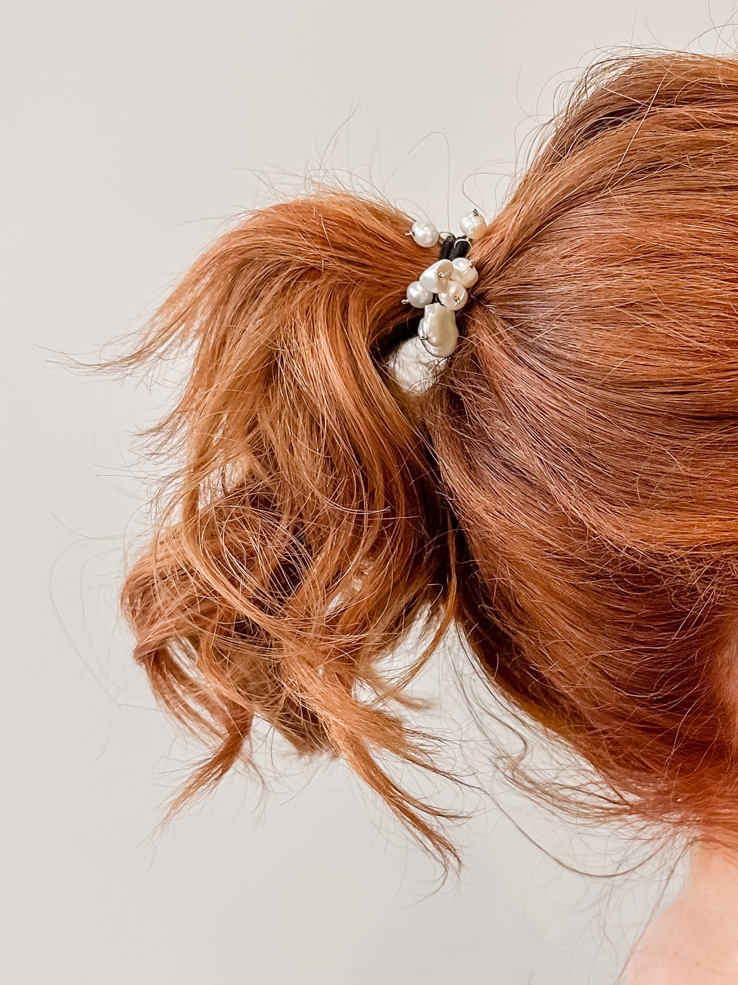 Elástico de cabelo para penteados e rabo de cavalo em tom preto com 8 perolas penduradas de diversos tamanhoas e formatos fixados com fio de prata prendendo cabelo de uma mulher ruiva com fundo branco.