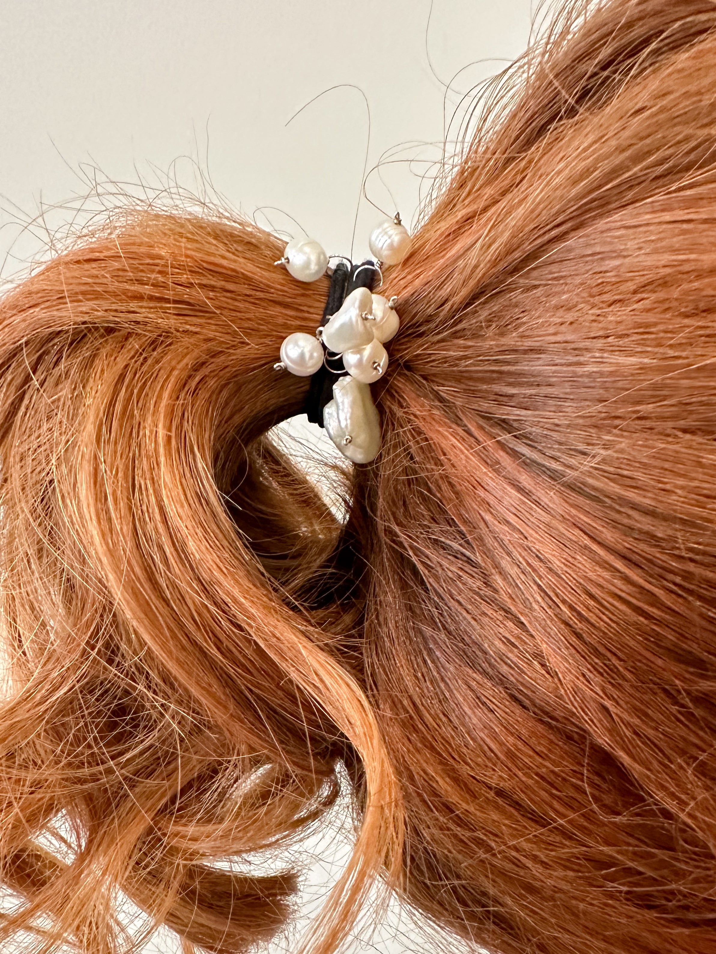Elástico de cabelo para penteados e rabo de cavalo em tom preto com 8 perolas penduradas de diversos tamanhoas e formatos fixados com fio de prata prendendo cabelo de uma mulher ruiva com fundo branco.