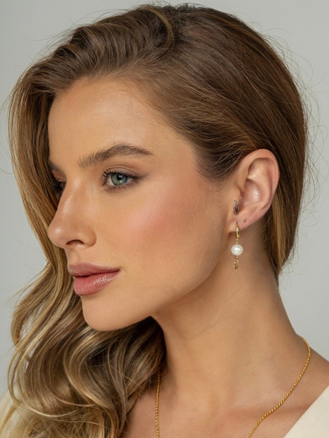 Mulher veste Brinco com uma mini argola em click e uma perola natural redonda pendurada e na ponta uma plaquinha redonda em prata 925 com banho de ouro 18k.